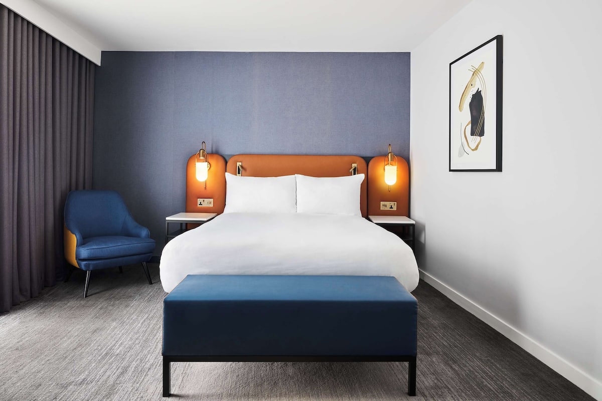 A hotel room with a large bed, two bedside tables with lamps, a blue chair, and a framed picture on the wall.