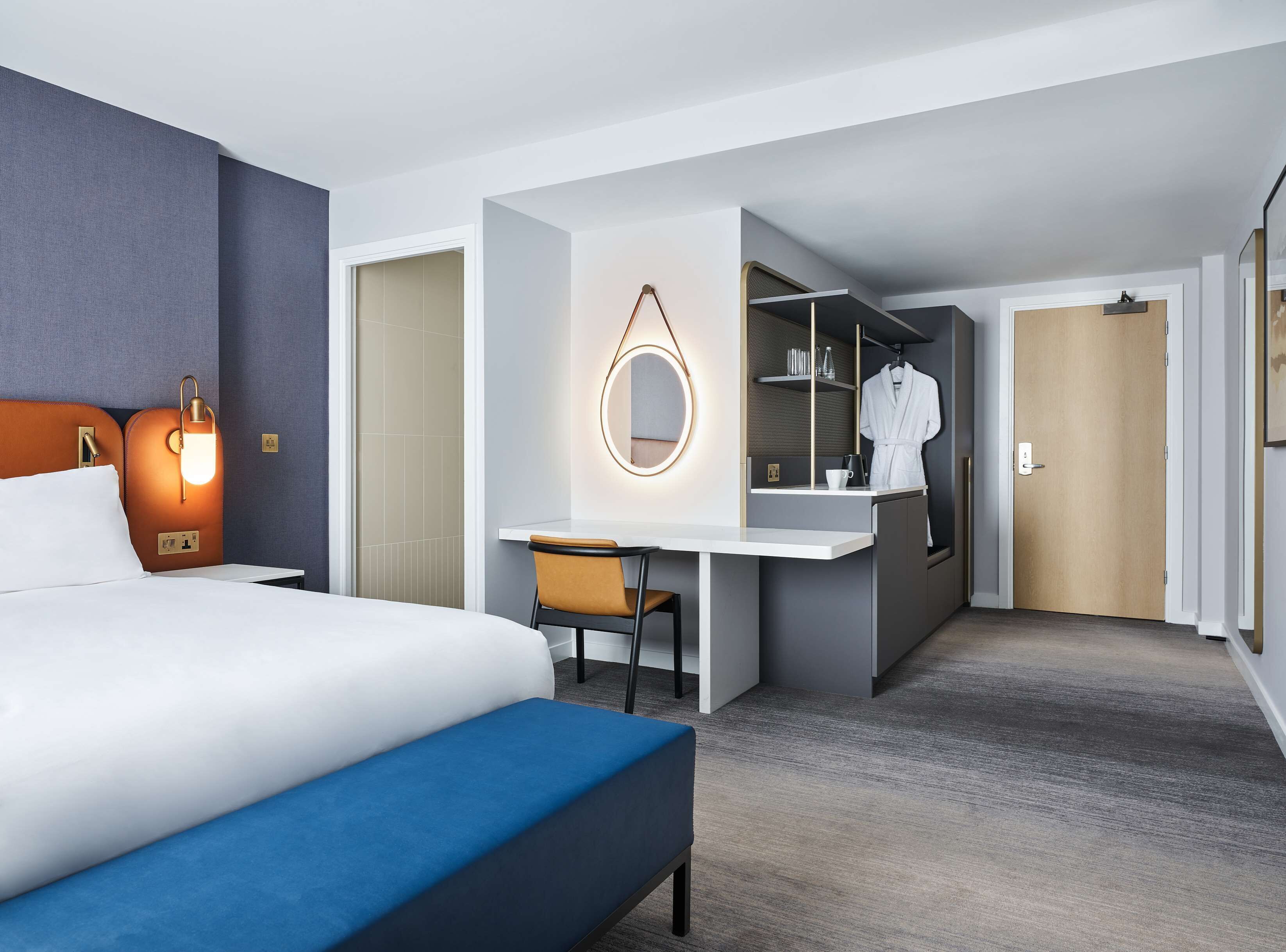 A hotel room with a bed, a desk, a chair, a robe, and a mirror.