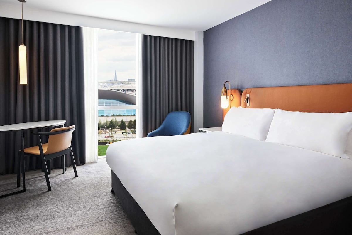 A hotel room with a large bed, a desk, a chair, and a view of a cityscape through the window.