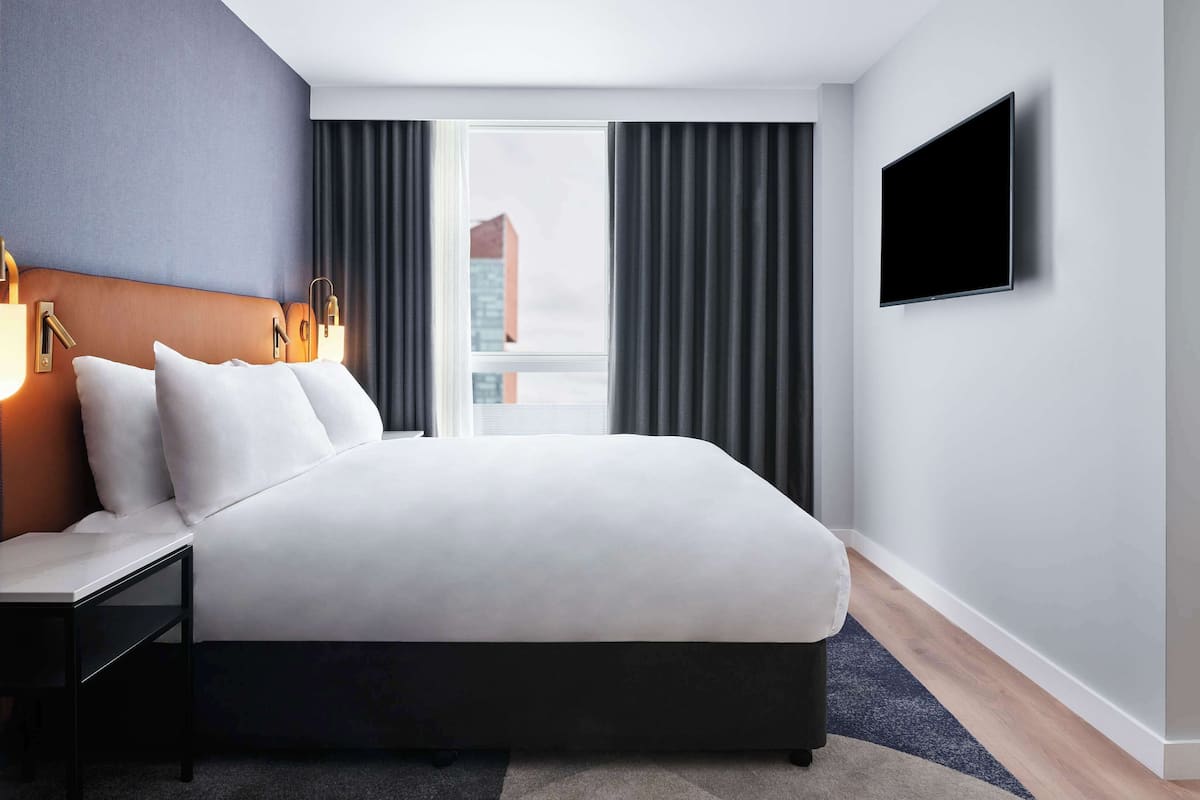 A modern hotel room with a large bed, a bedside table, a flat-screen TV, and a view of a building through the window.