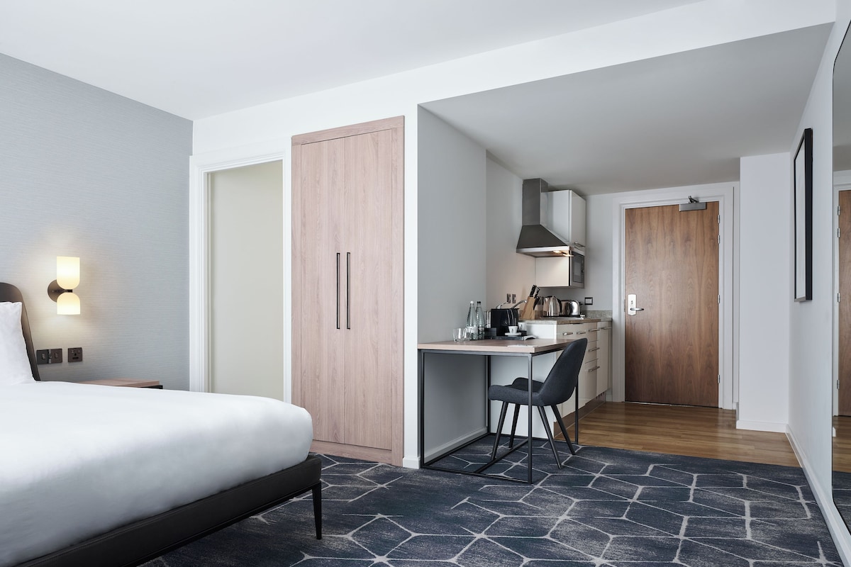 A hotel room with a bed, a desk, a chair, a kitchenette, and a wardrobe.