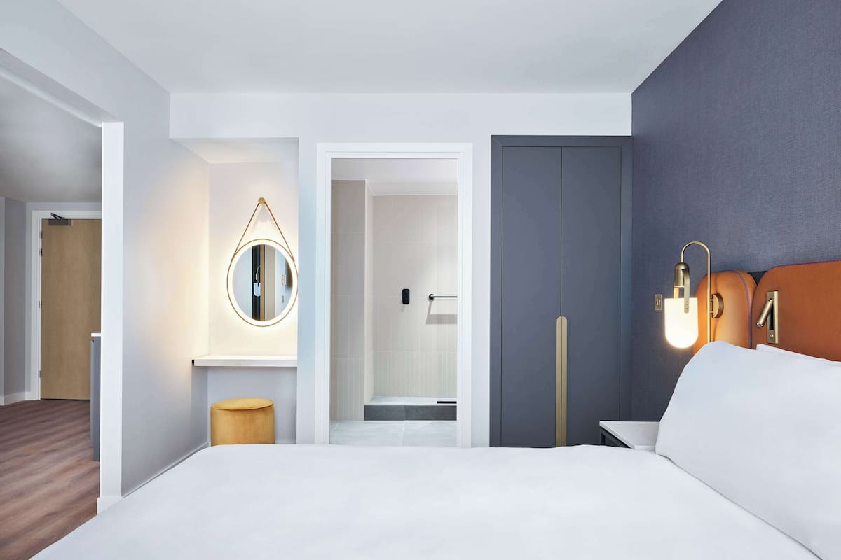 A modern hotel room with a large bed, a round mirror, a bedside table, and a nightstand.