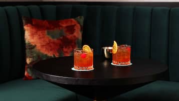 Two cocktails on a round table with a dark surface, set against a green upholstered sofa with a colorful pillow.