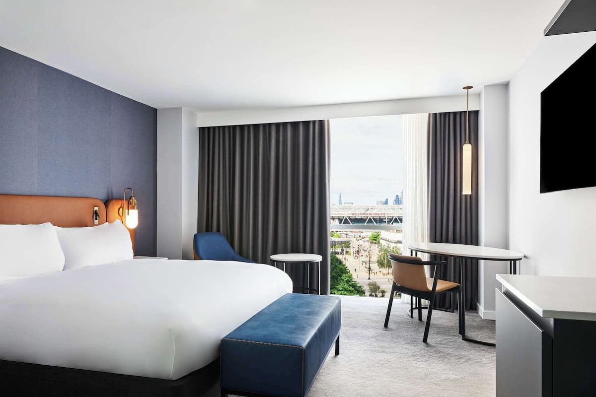 A modern hotel room with a large bed, a desk, a chair, and a view of a cityscape.