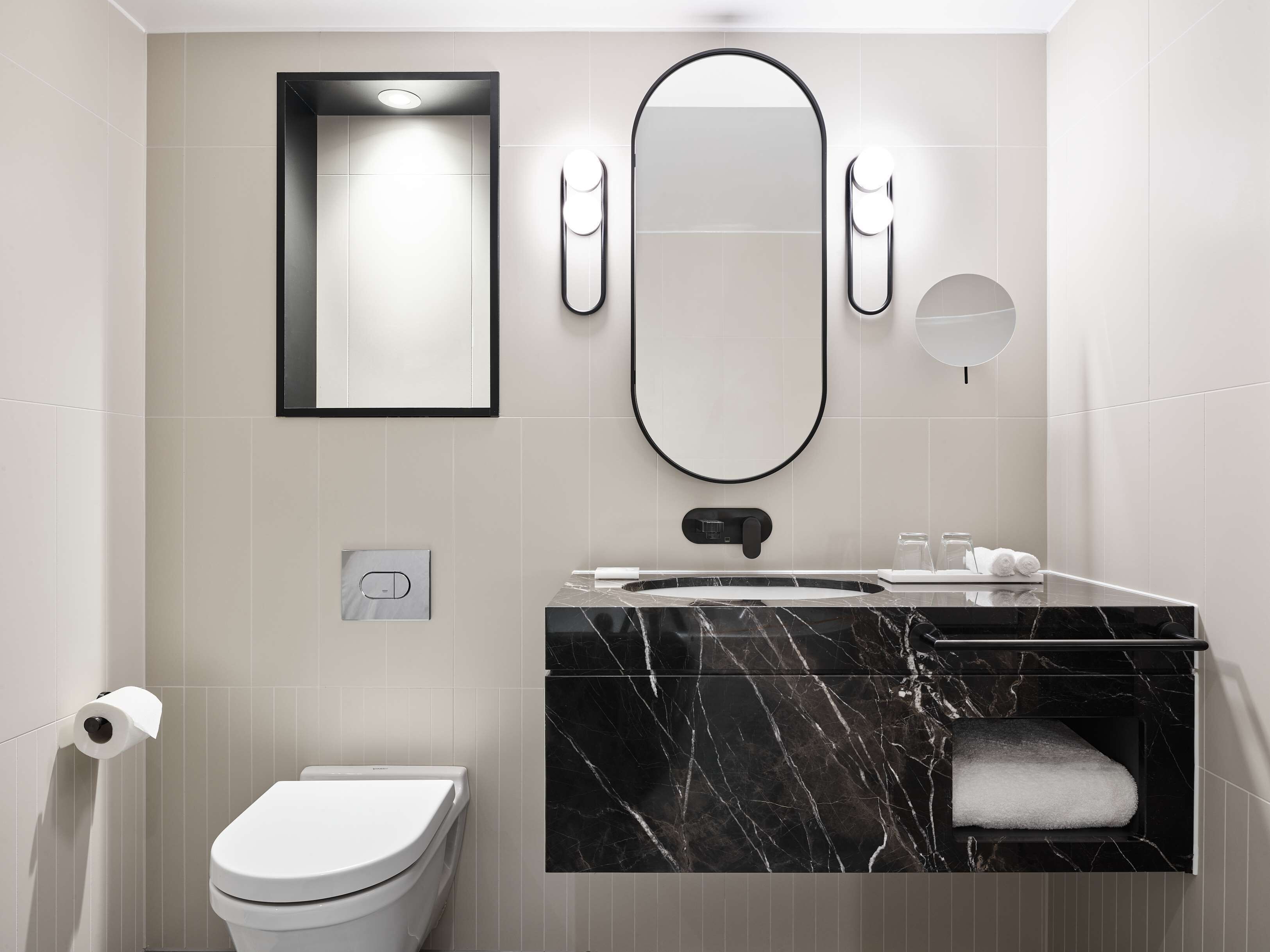 A modern bathroom with a toilet, a sink with a marble countertop, a large mirror, and two wall-mounted lights.