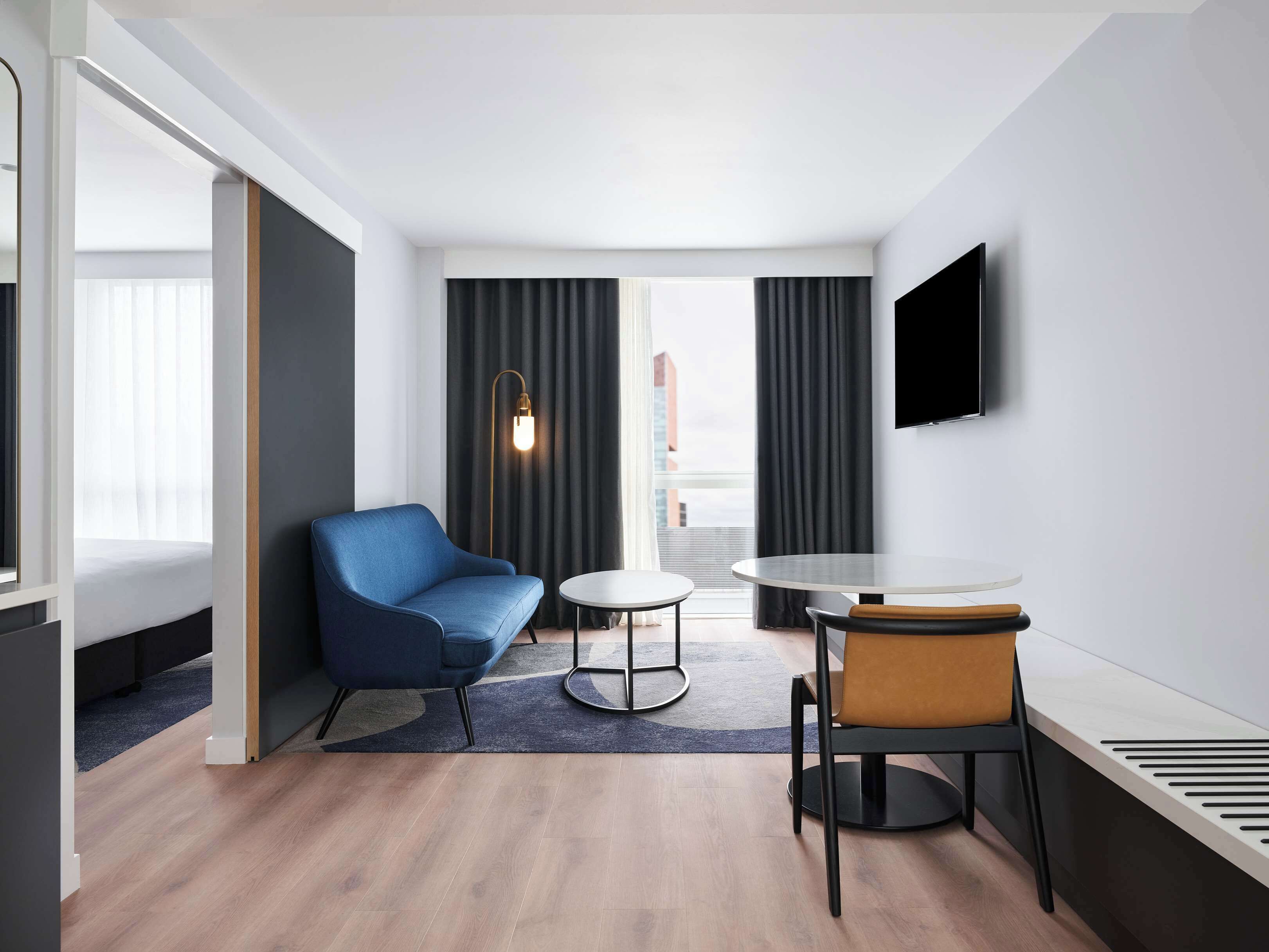 Una habitación de hotel moderna con un sillón azul, una mesa redonda, un sillón de cuero marrón, un televisor de pantalla plana montado en la pared y un ventanal con cortinas.