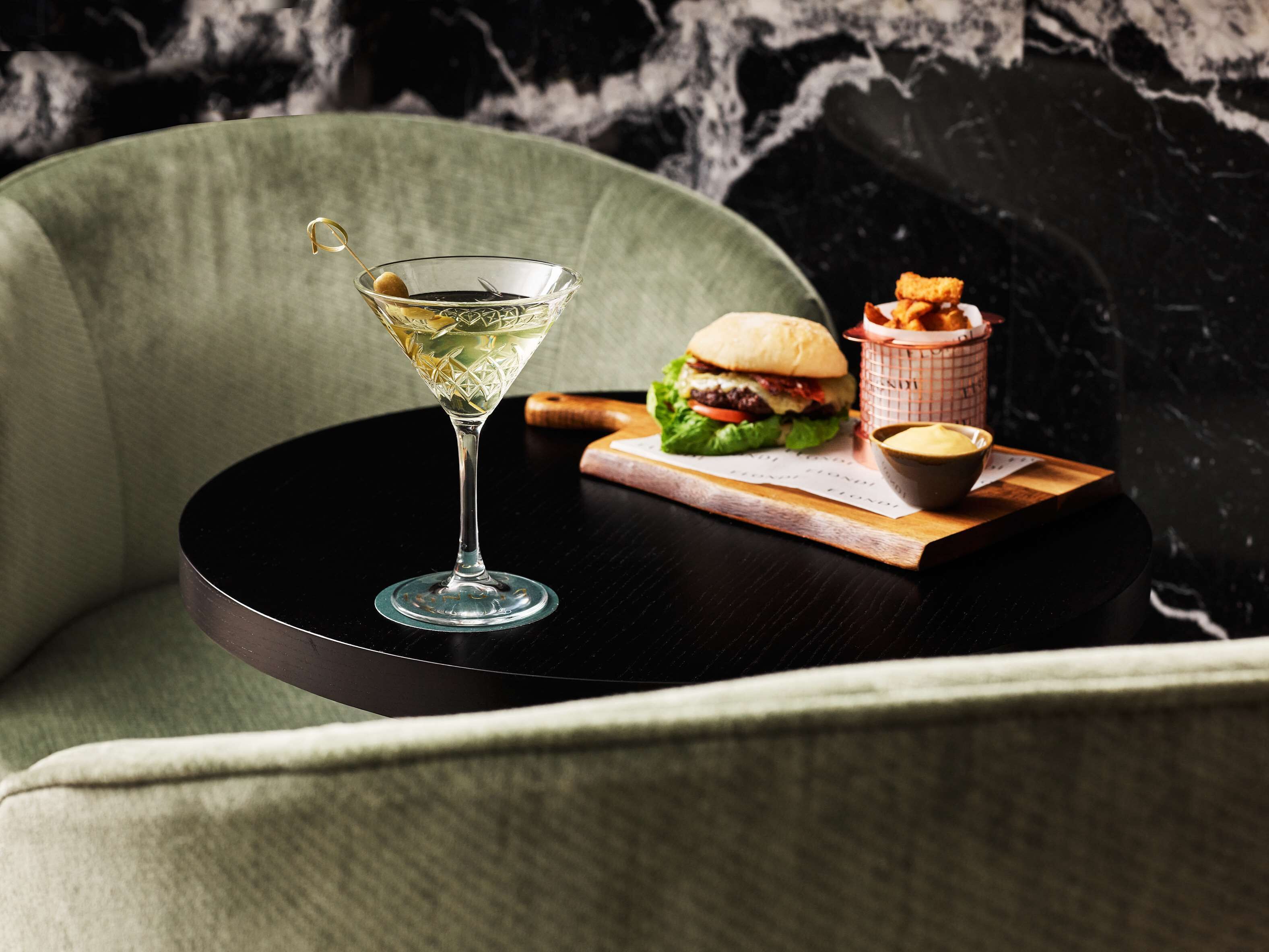 A martini glass with a lemon twist on a black coaster, a burger, and a basket of fries on a wooden tray.