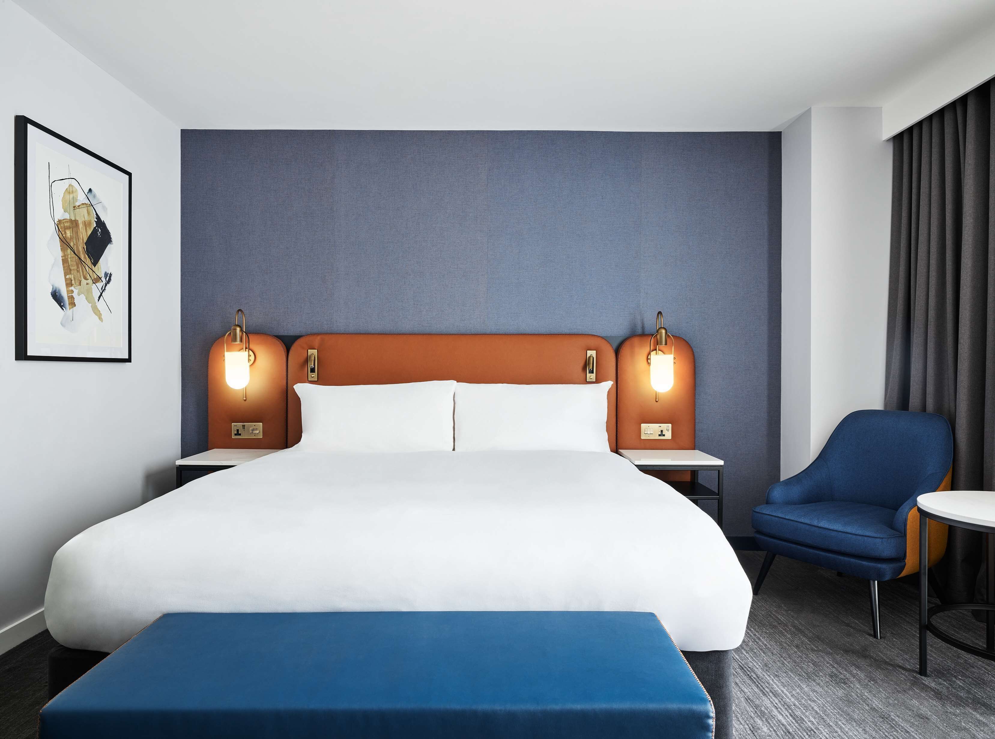 A hotel room with a large bed, two bedside tables, a chair, and a blue bench.