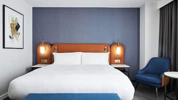 Quarto de hotel com uma cama grande, duas mesas de cabeceira, uma cadeira e um banco azul.