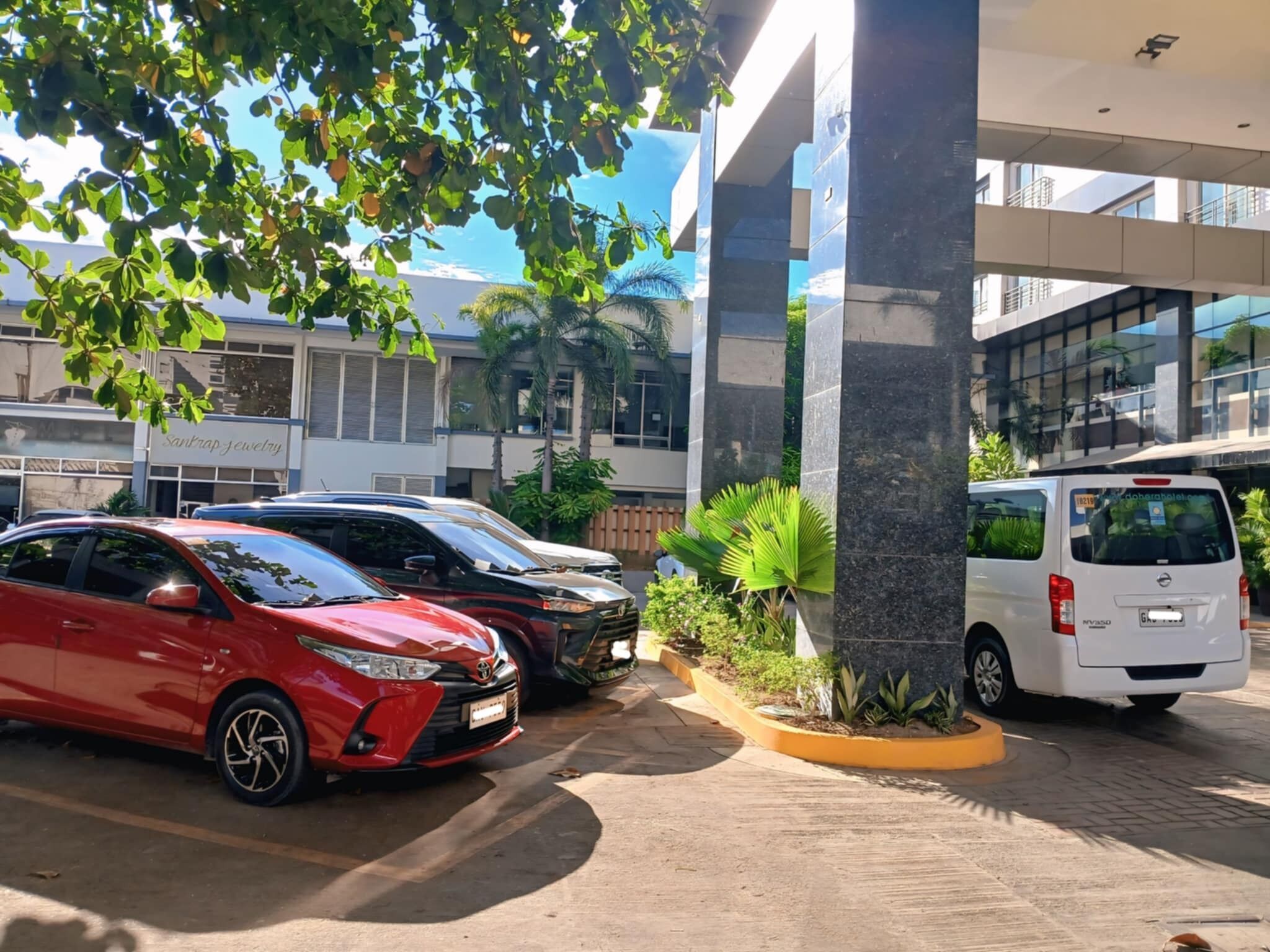 A parking area with several cars, a building with a modern design, and a tree providing shade.