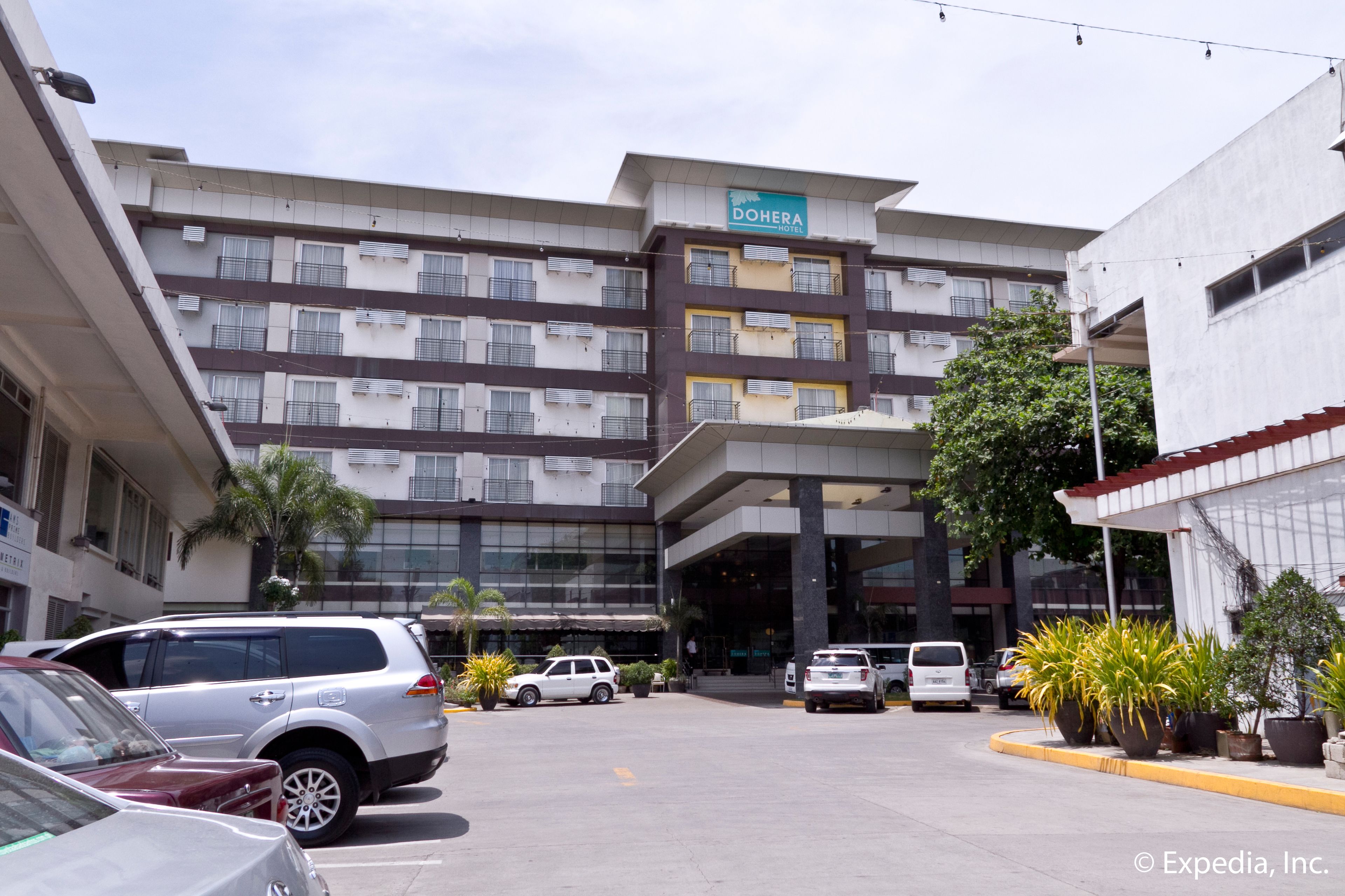 Un hotel de varios pisos con un diseño moderno, que destaca un letrero DOHERA, un estacionamiento con varios coches y plantas en macetas a lo largo de la acera.