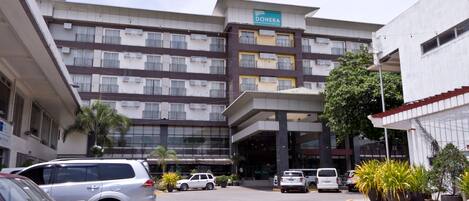 Un hotel de varios pisos con un diseño moderno, que destaca un letrero DOHERA, un estacionamiento con varios coches y plantas en macetas a lo largo de la acera.