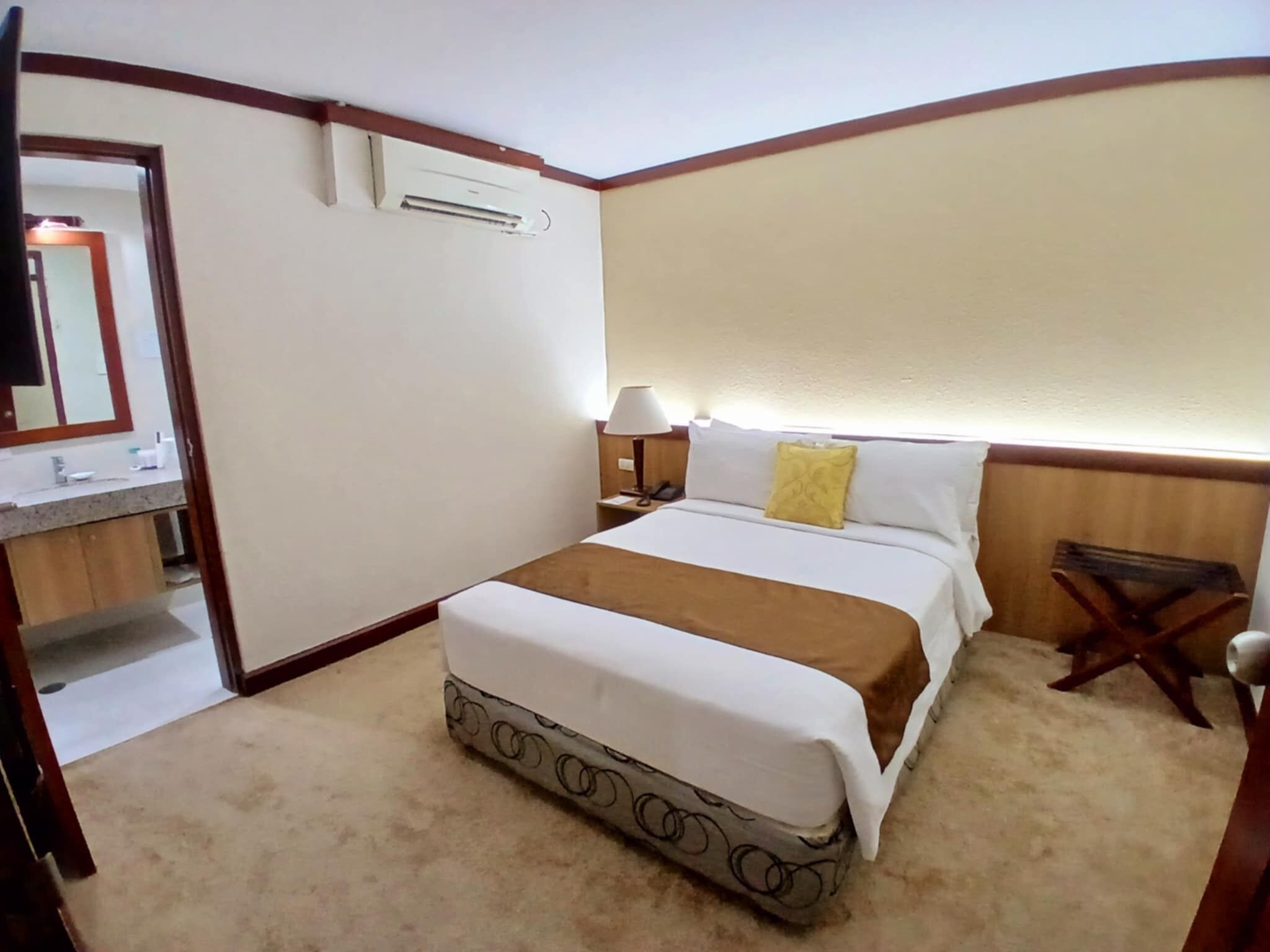 A hotel room with a bed, a bedside table with a lamp, a mirror, and an air conditioning unit.