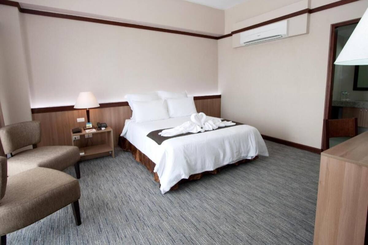 A hotel room with a bed, a chair, a nightstand, a lamp, and an air conditioning unit.