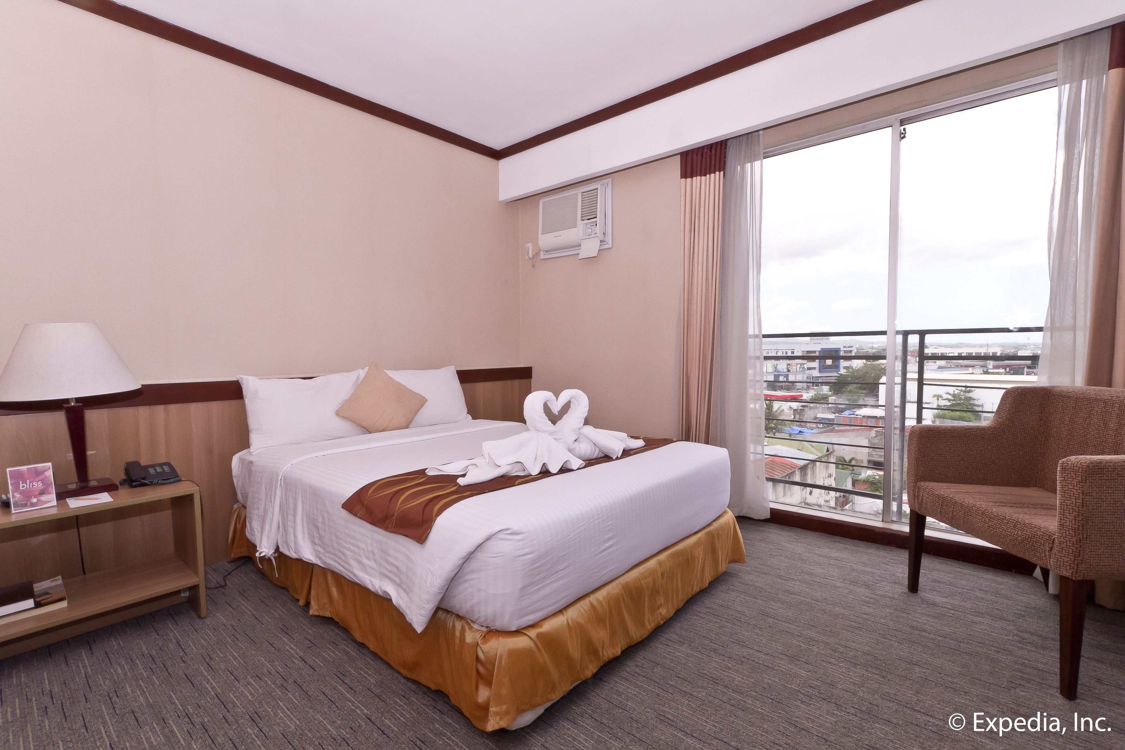 Habitación de hotel con una cama grande, una mesita de noche, una silla y vistas de la ciudad a través de un ventanal.