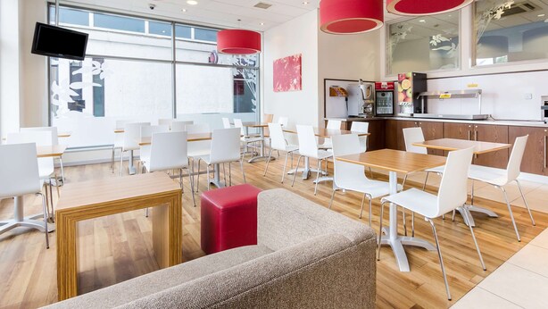 A modern cafe with white chairs, wooden tables, and red accents. - Travelodge Farnborough Central (Farnborough)