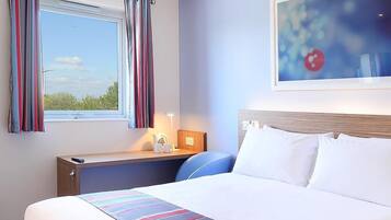 A hotel room with a bed, a desk, a window with curtains, a painting on the wall, and a view of trees outside.