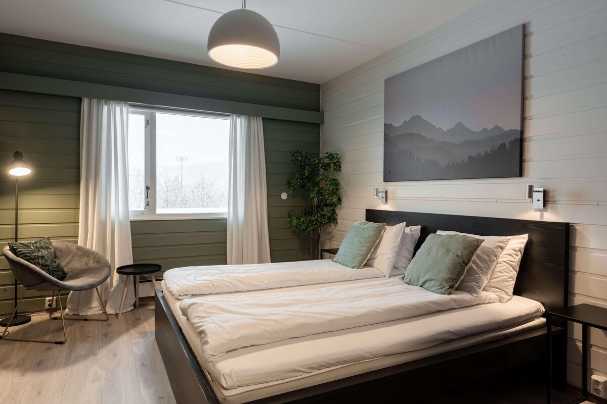 Ein Doppelbett mit weißer Bettwäsche, ein Sessel, ein kleiner Tisch, eine Pendelleuchte, ein Fenster mit Vorhängen, eine Pflanze und ein Gemälde an der Wand.