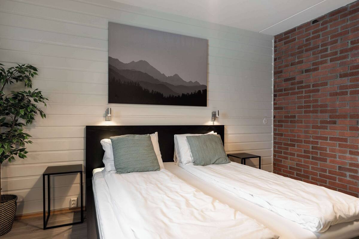 Une chambre avec deux lits, un tableau accroché au mur, une plante et un mur en briques.