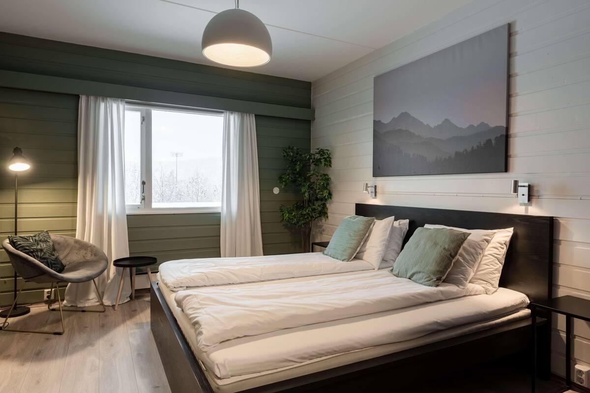 Un lit double avec une literie blanche, une chaise, une petite table, un luminaire suspendu, une fenêtre avec des rideaux, une plante et un tableau au mur.