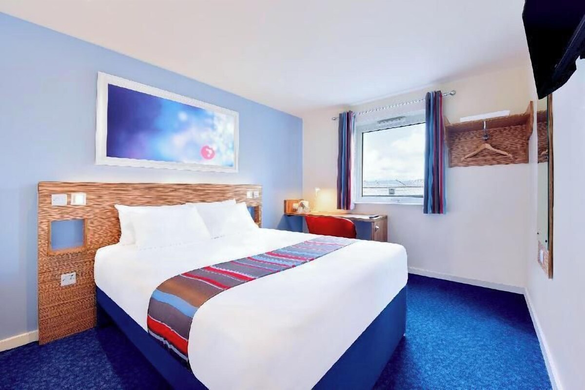 Une chambre d’hôtel avec un grand lit, un téléviseur fixé au mur, une fenêtre avec des rideaux et une petite table avec une lampe.