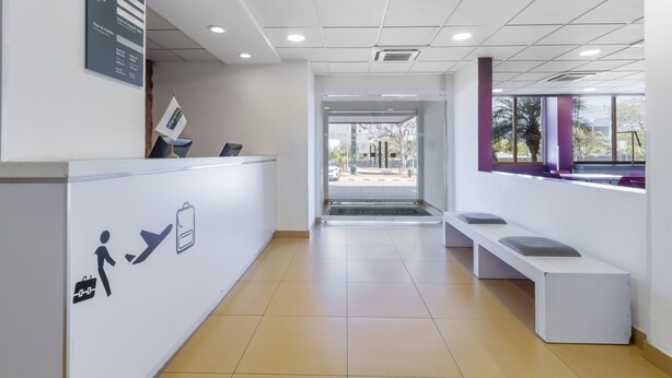 Zona de recepción con mostrador blanco, área de espera con bancos y vista al exterior a través de amplias ventanas.
