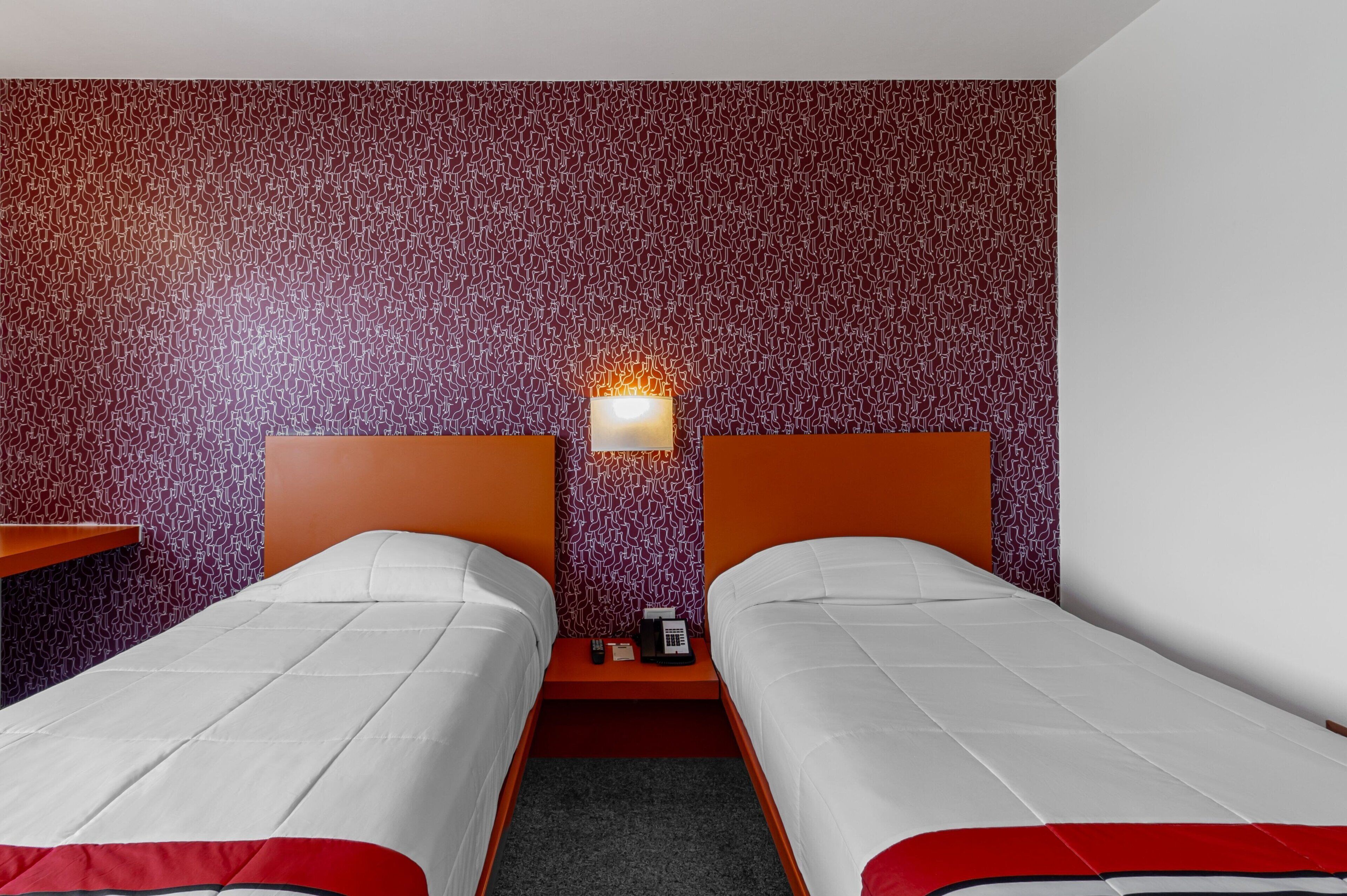A hotel room with two beds, a nightstand with a lamp, and a patterned wall.