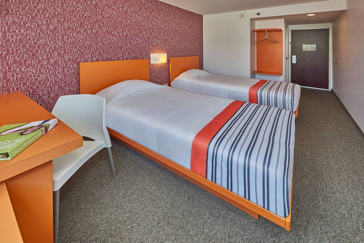 Une chambre d’hôtel avec deux lits, un bureau et une chaise. Un mur est décoré d’un motif et la porte porte une pancarte.