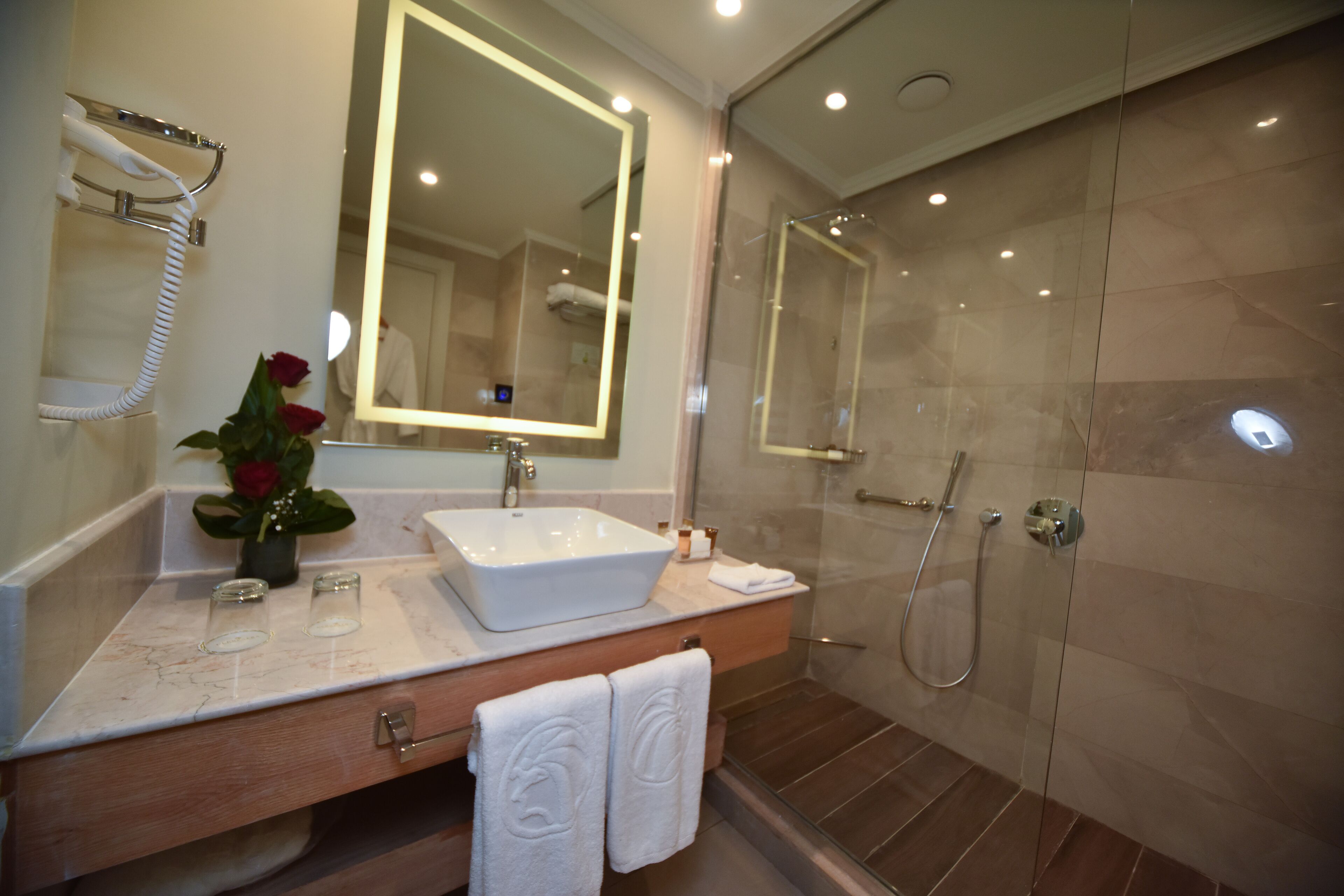 Un baño moderno con un espejo grande, un lavamanos blanco, una ducha con mampara de vidrio y un mueble de tocador con toallero.