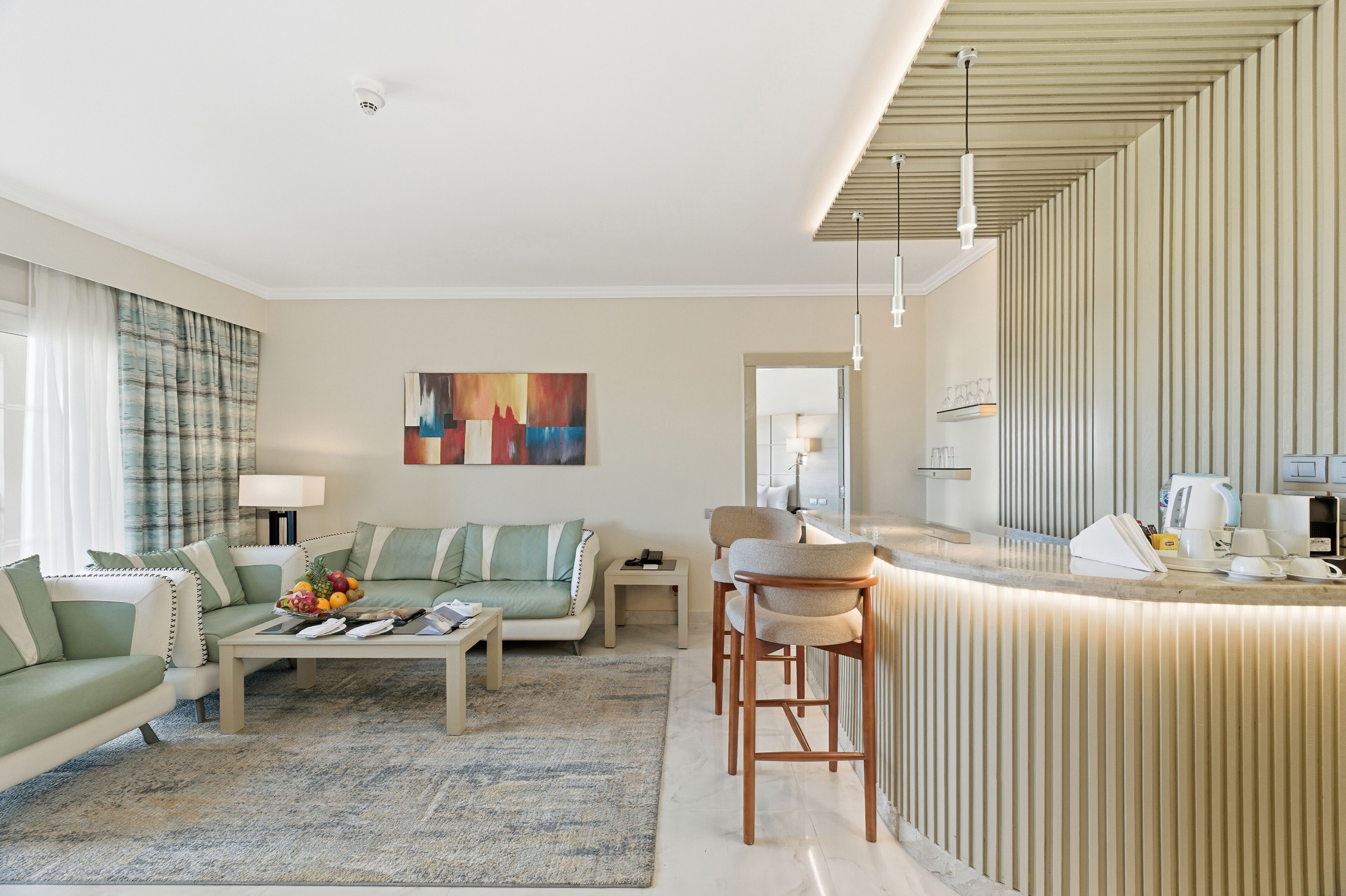 Una sala moderna con sofá, mesa de centro y sillas, una zona de cocina con encimera y electrodomésticos, y un cuadro abstracto grande en la pared.