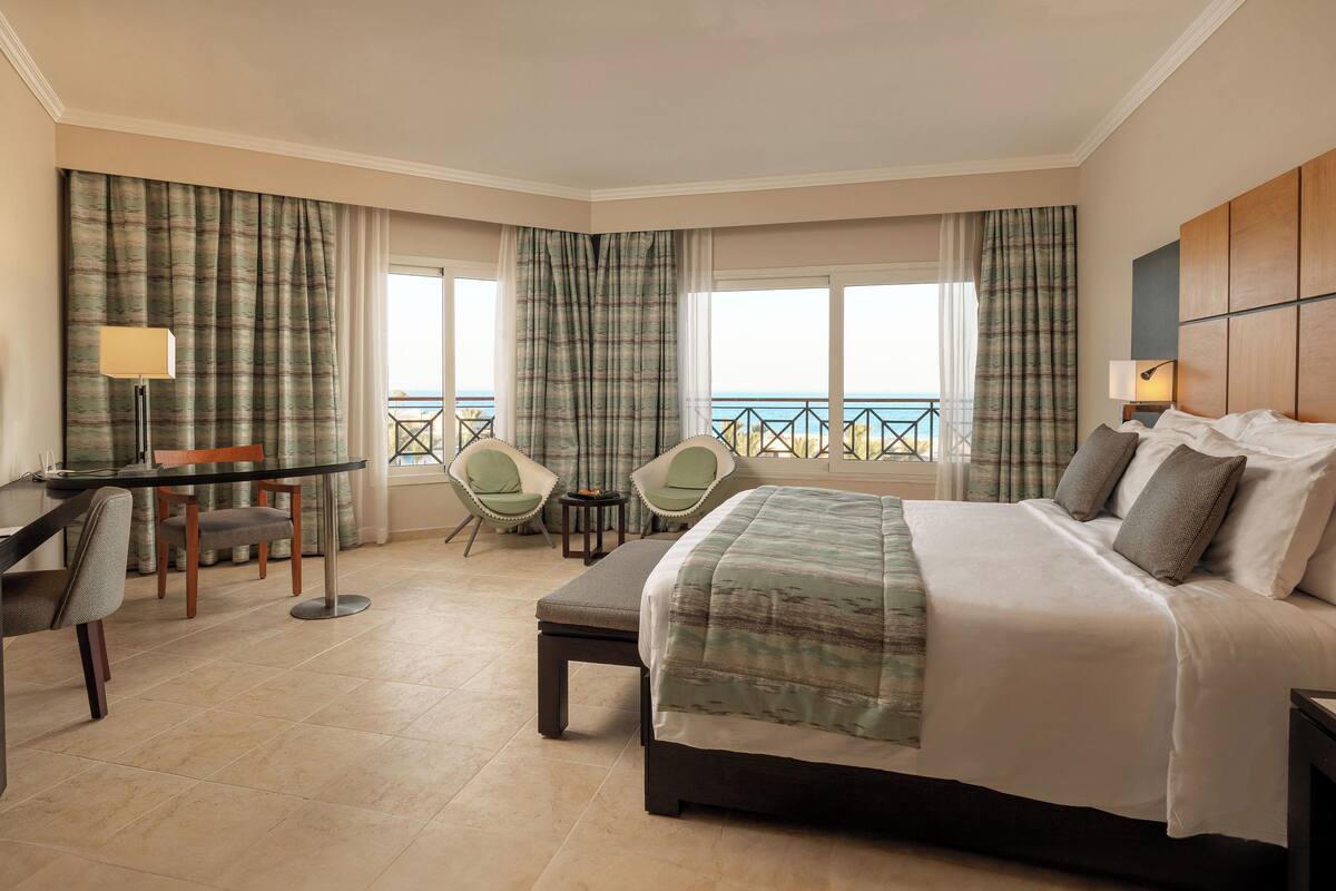 Une chambre d’hôtel comprenant un lit, un bureau, deux chaises, une petite table et de grandes fenêtres donnant sur la mer.