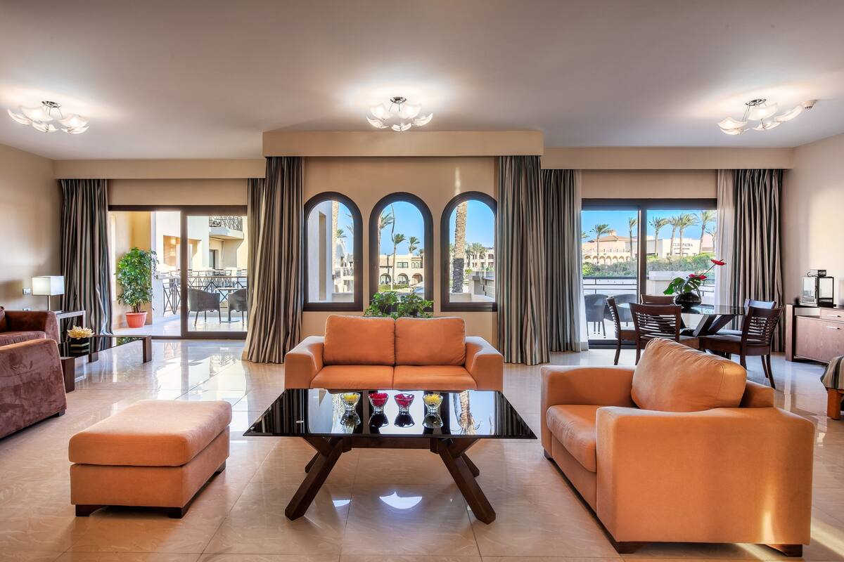 Un salon spacieux comprenant un canapé, un pouf et une table basse en verre. De grandes fenêtres, ornées de rideaux, offrent une vue sur l’extérieur.