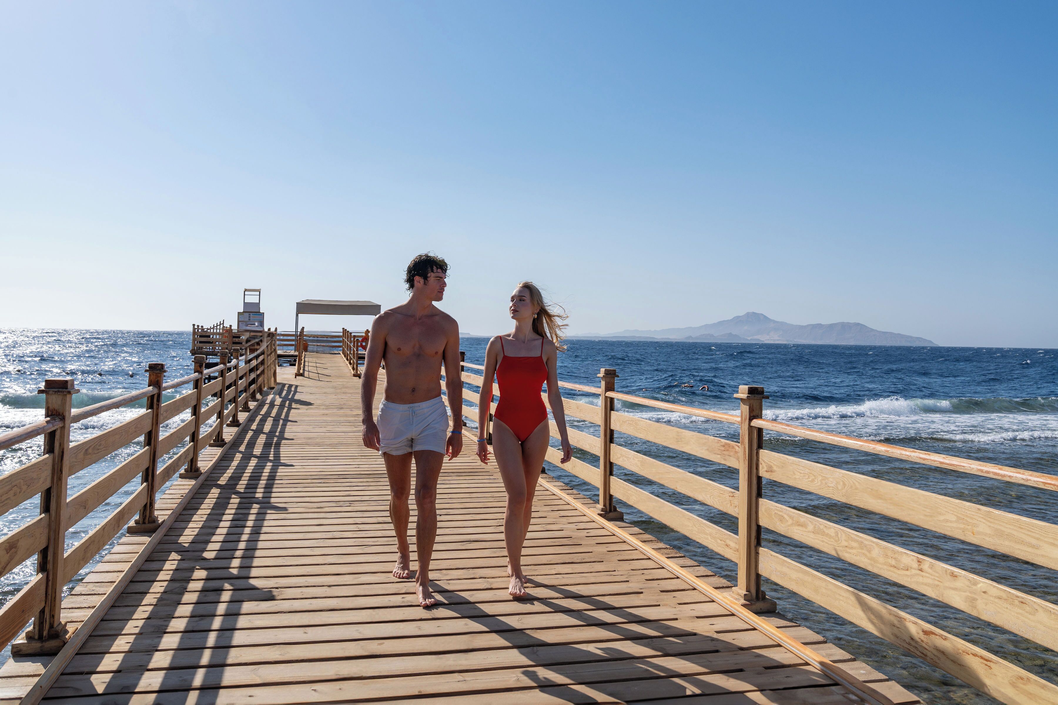 A wooden pier extending into the sea with a clear sky and distant mountains.