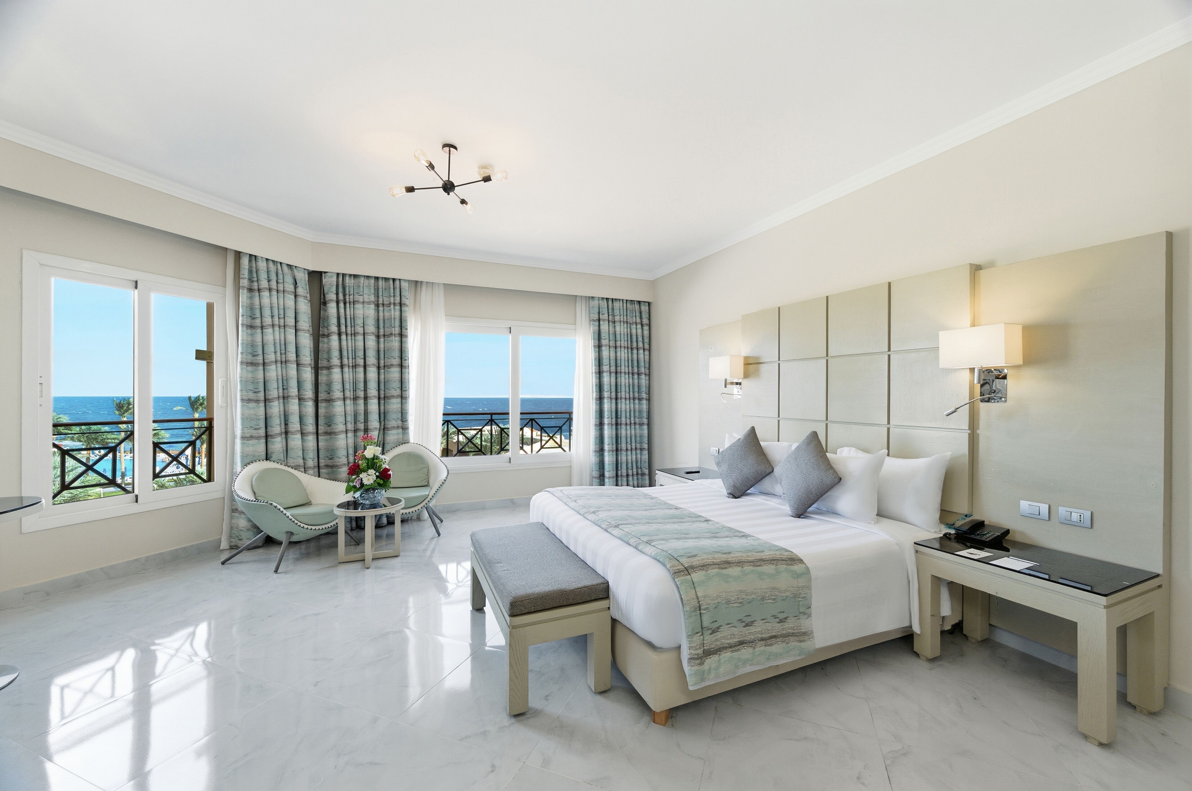 A spacious bedroom with a large bed, bedside tables, a desk, and a view of the sea.