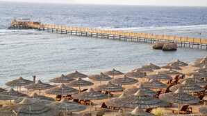 Een strand met een houten pier die zich uitstrekt in de zee, omringd door rieten parasols en strandstoelen.