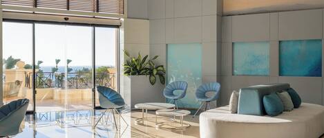 A modern lounge with a glass floor, blue chairs, a white sofa, and artwork on the walls.