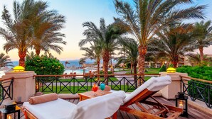 Une terrasse en bois avec des chaises longues, une table avec des boissons et une vue sur la mer et les palmiers.