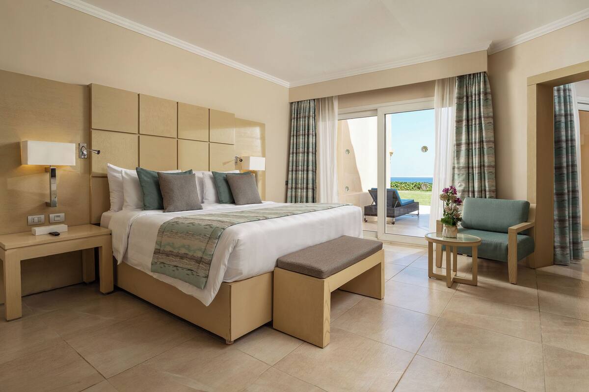 Une chambre d’hôtel moderne avec un grand lit, une table de chevet avec une lampe, un banc au pied du lit, une petite table ronde ornée d’un vase de fleurs et une vue sur l’extérieur grâce à une porte-fenêtre coulissante.