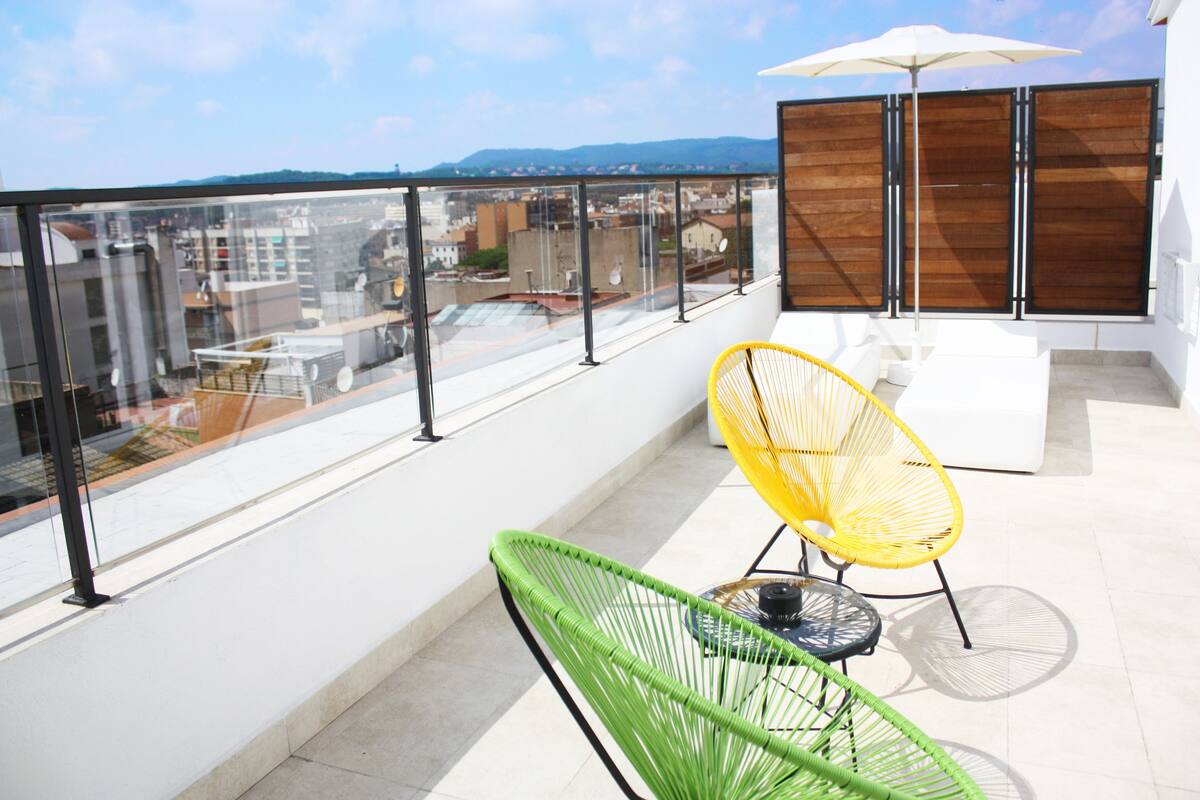 Un balcon doté d’une rambarde en verre, une chaise jaune, une chaise verte, une table blanche et une cloison en bois.