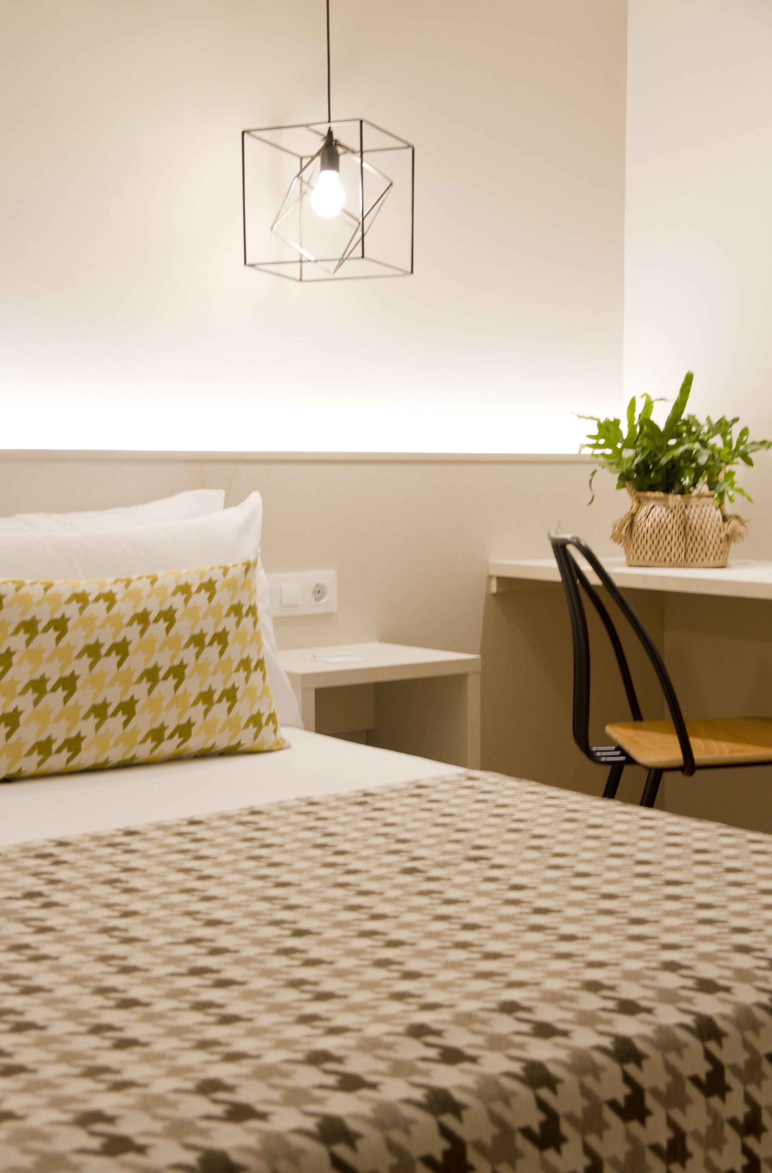 A hotel room with a bed, a desk, a chair, a hanging light fixture, and a potted plant.