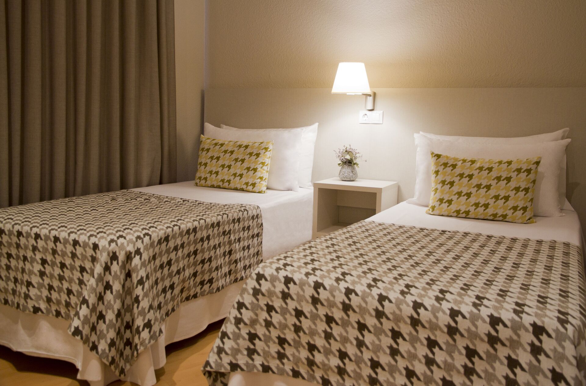 Ett hotellrum med två sängar, var och en med ett mönstrat överkast och matchande kuddar. Mellan sängarna står ett nattduksbord med en lampa.