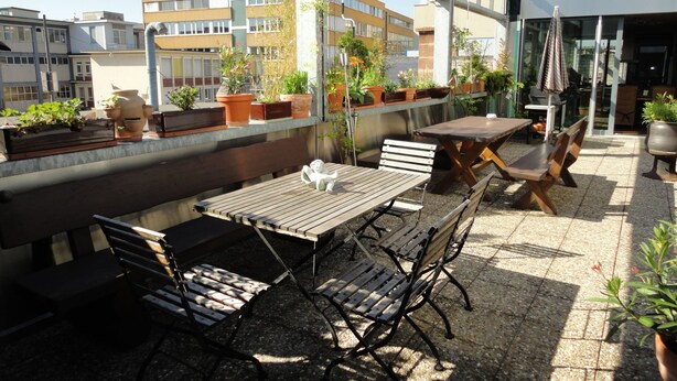 Une terrasse sur le toit, avec des tables et des chaises en bois, des plantes en pot et une vue sur les bâtiments en arrière-plan.