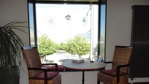 Une pièce avec deux fauteuils en osier disposés autour d’une table ronde, une plante en pot et une vue sur une rue et des arbres, visible à travers une grande fenêtre.