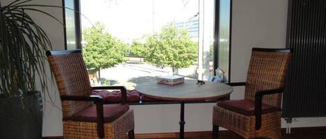 A room with two wicker chairs around a round table, a potted plant, and a view of a street and trees through a large window.