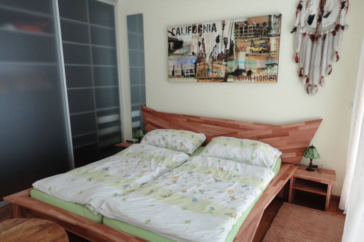 A bedroom with a wooden bed, two pillows, a nightstand with a lamp, a wall art piece, and a sliding door.