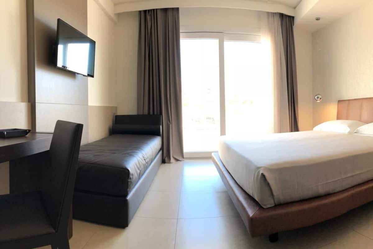 A hotel room with a bed, a desk, a chair, a TV mounted on the wall, and a sliding door leading to a balcony.
