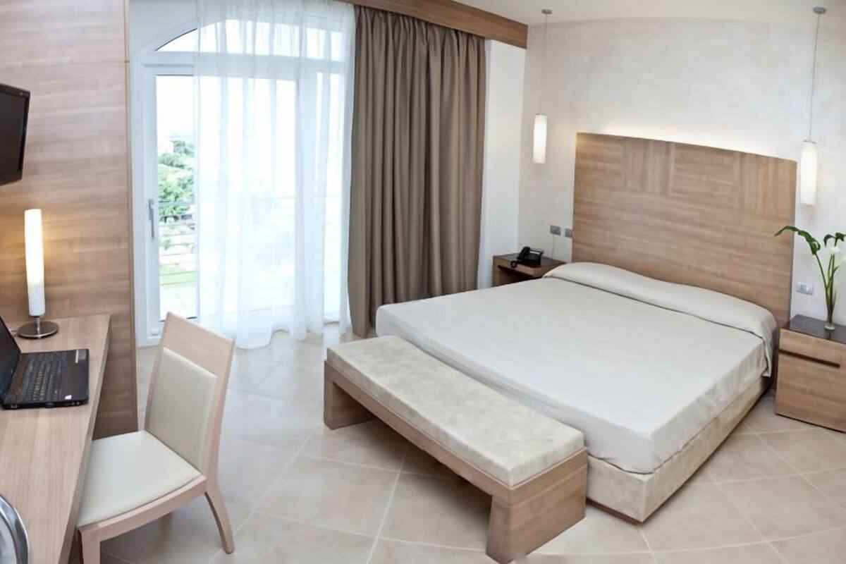 A hotel room with a bed, a desk with a laptop, a chair, a bench, a TV, a nightstand, and a window with curtains.