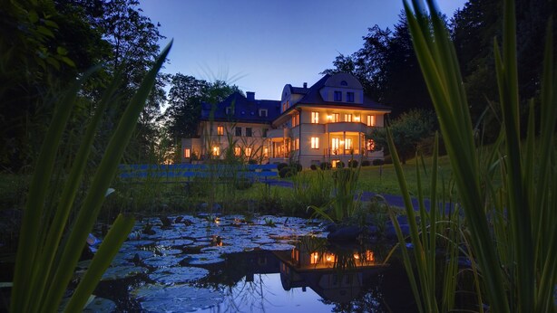 A large, illuminated house with a pond in the foreground. - hideauts hotels Der Rosengarten. (Fuessen)
