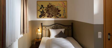 A single bed with a dark frame, white bedding, a bedside table with a lamp, a radiator, a painting above the bed, and a door to the right.