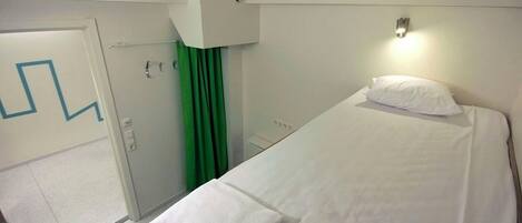 A hotel room with a single bed, a white headboard, a bedside lamp, and a green curtain.